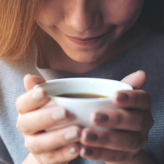 A woman with long hair smiles gently while holding a steaming white cup of coffee close to her face, suggesting enjoyment in a cozy indoor setting.