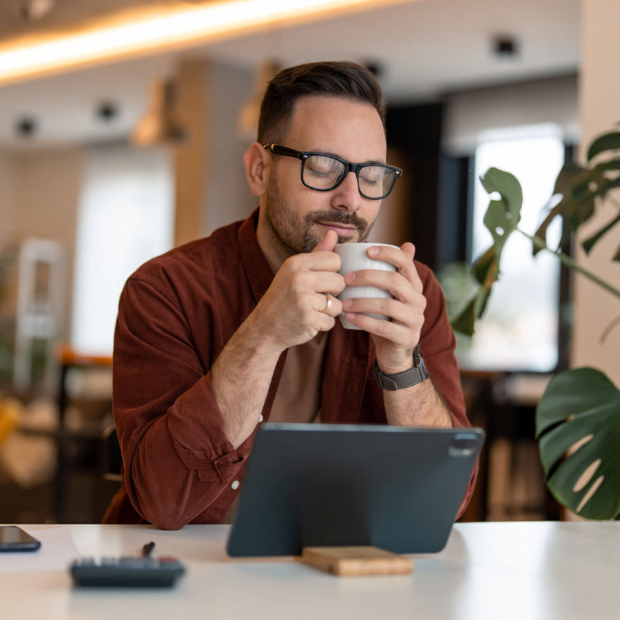A man with glasses sits at a table in a cozy café, holding a cup of coffee with his eyes closed, enjoying the moment, while a tablet and a plant are visible nearby.