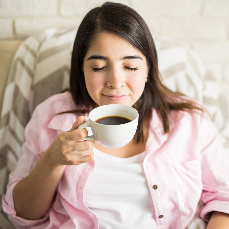 A woman with closed eyes enjoys a cup of coffee while sitting comfortably on a couch, wearing a pink shirt and surrounded by a cozy blanket and a light-colored wall.