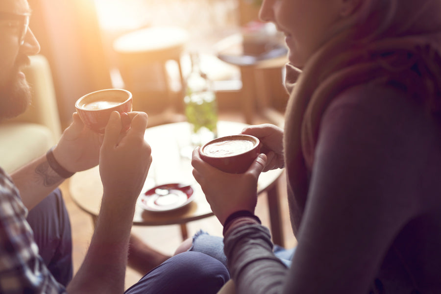 A man and woman sit close together, smiling and holding steaming cups of coffee in a cozy café with warm sunlight streaming through the window.