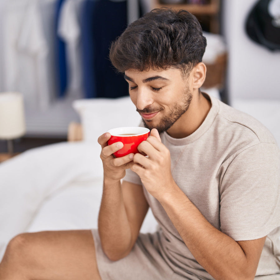 A young man with short dark hair sits on a bed, holding a red bowl close to his face with a content smile, enjoying a moment of relaxation in a cozy bedroom setting.