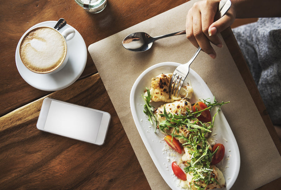 A hand holds a fork while preparing to eat a plate of cheesy pasta topped with arugula and tomatoes, alongside a cup of cappuccino and a smartphone on a wooden table.