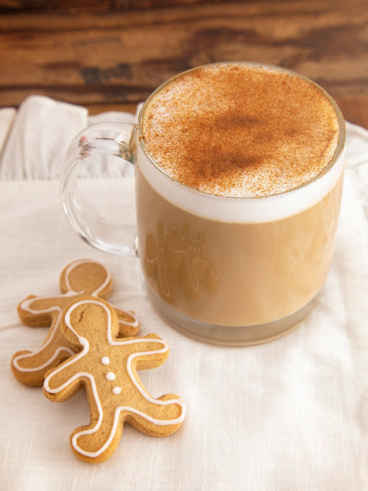A clear glass mug filled with frothy coffee topped with cinnamon sits on a light fabric, accompanied by two gingerbread cookies shaped like little people, adding a festive touch.