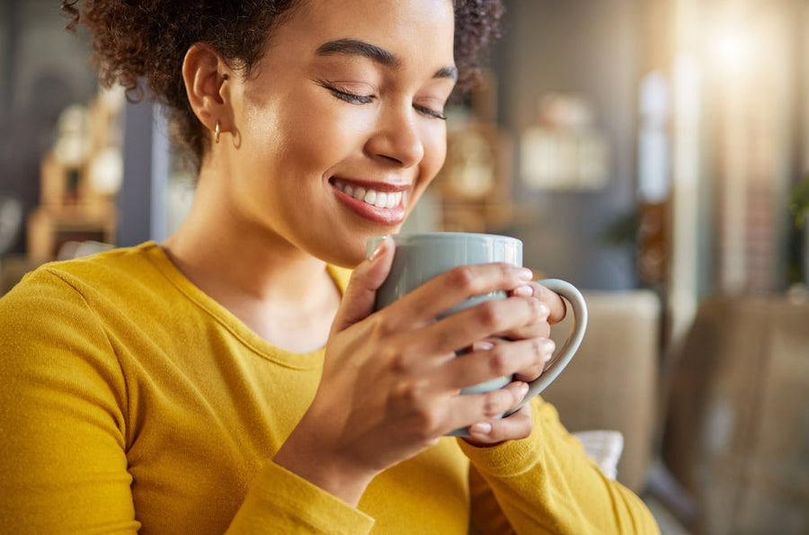 A young woman in a yellow sweater smiles with her eyes closed while holding a light blue mug, enjoying a moment of warmth in a cozy café setting.