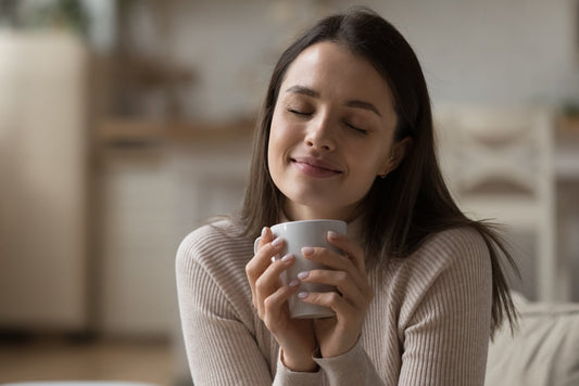 A young woman with long brown hair smiles gently while holding a white mug close to her face, enjoying a moment of warmth and comfort in a cozy, softly lit kitchen.