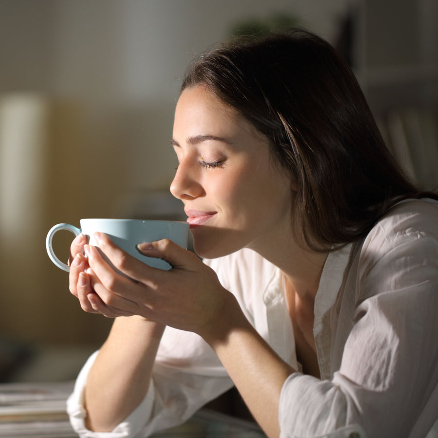 A woman in a white shirt gently holds a light blue cup close to her face, eyes closed and smiling, in a softly lit indoor setting, enjoying a moment of warmth and comfort.