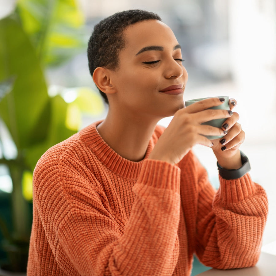 A woman in an orange sweater smiles contentedly while holding a teal cup, sitting in a bright café with green plants in the background.