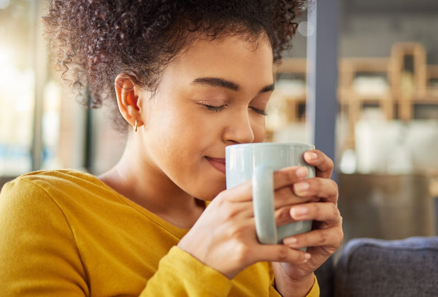 A young woman with curly hair closes her eyes and smiles while holding a light blue mug close to her face in a cozy cafe setting.