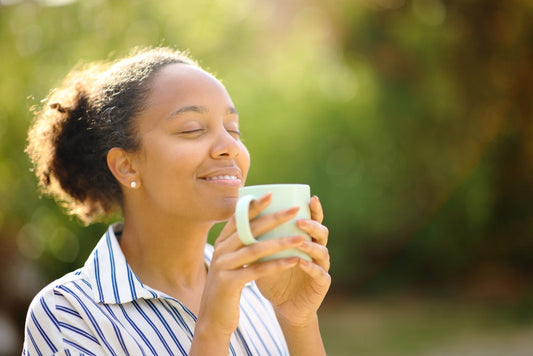 A woman with curly hair smiles gently while holding a light blue mug close to her face, enjoying the aroma, with a blurred green background suggesting a sunny outdoor setting.