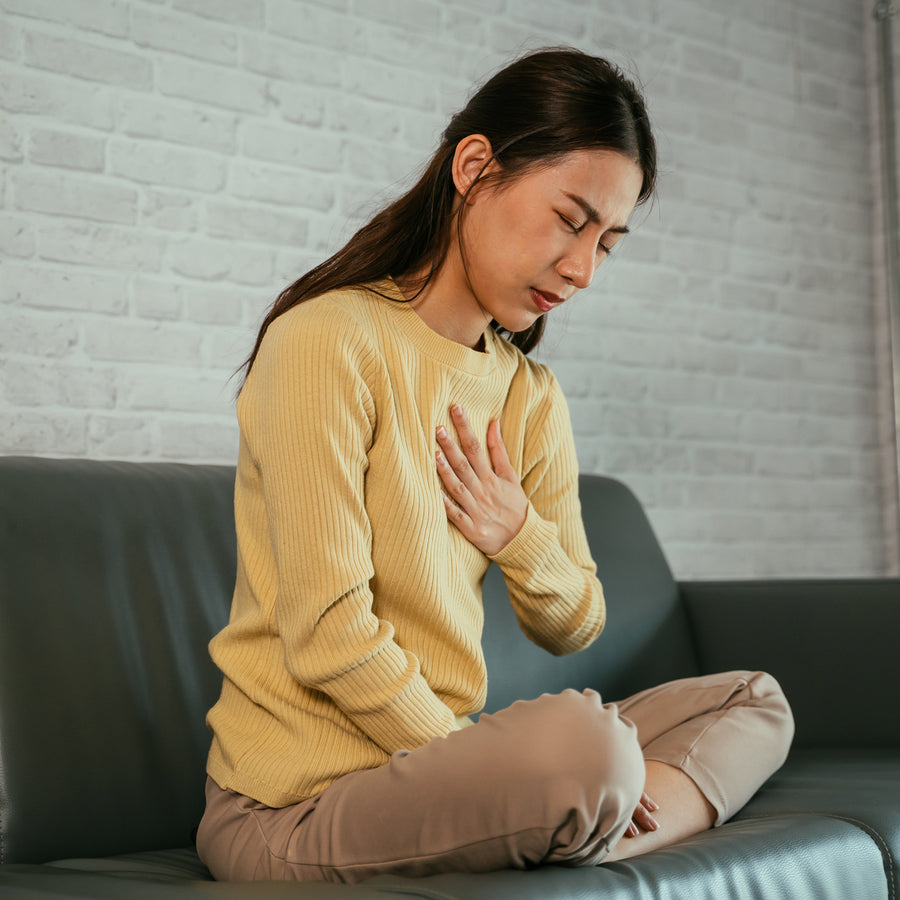 A woman in a yellow sweater sits on a couch, holding her chest with a pained expression, suggesting she is experiencing discomfort or distress in a modern, light-colored room.