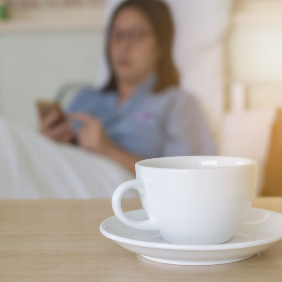 A white coffee cup sits on a saucer in the foreground, while a person in a blue shirt is blurred in the background, focused on their phone while resting in bed.