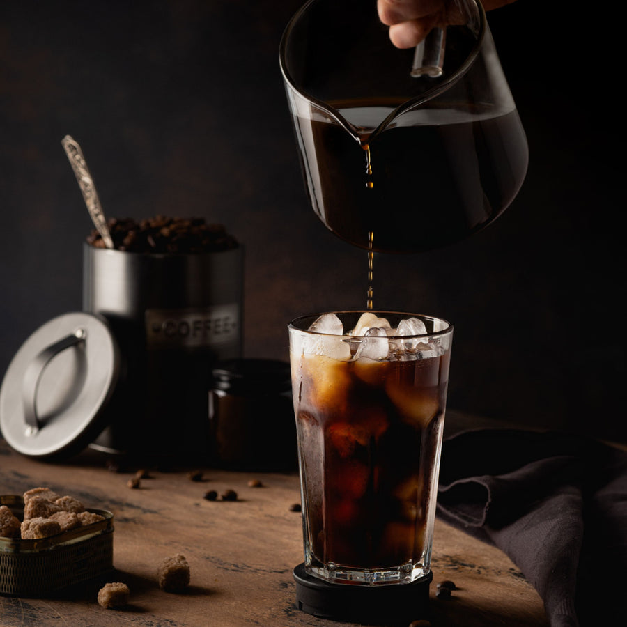 A hand pours dark coffee from a glass pitcher into a tall glass filled with ice, surrounded by coffee beans, a sugar container, and a dark cloth on a wooden table.