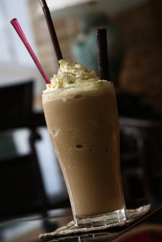 A tall glass of iced coffee topped with whipped cream and chocolate shavings sits on a coaster, featuring two colorful straws and a dark chocolate stick, against a blurred café background.
