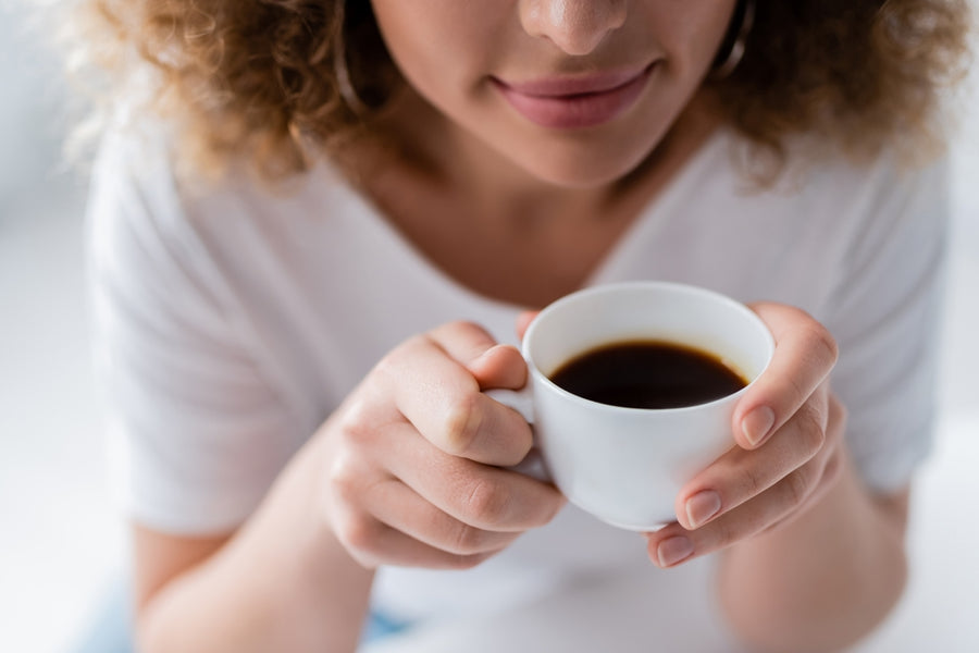 A woman with curly hair holds a small white cup of coffee close to her lips, smiling softly while sitting in a bright, cozy indoor setting.
