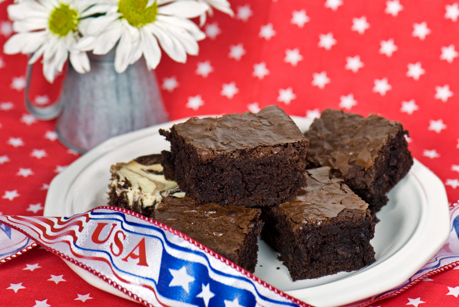 Brownies are arranged on a white plate with a decorative ribbon and a vase of daisies in the background, set against a red tablecloth adorned with white stars.