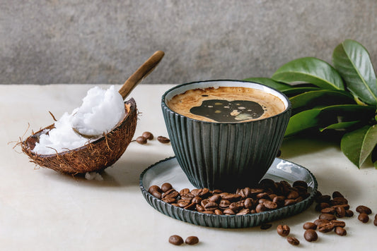 A dark coffee in a textured cup sits on a plate surrounded by coffee beans, next to a coconut shell filled with white coconut cream, with green leaves in the background.