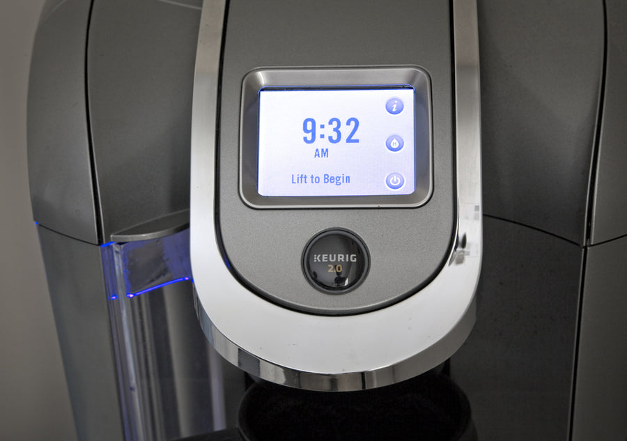 A close-up of a Keurig coffee maker's control panel displaying the time as 9:32 AM and a prompt that says "Lift to Begin," with a sleek silver design and blue lighting.