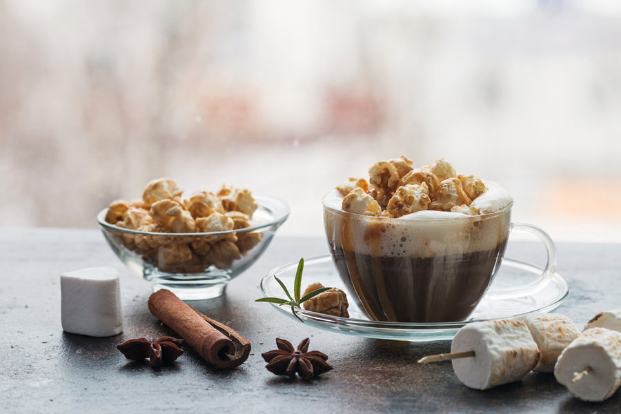 A glass cup filled with coffee topped with popcorn sits next to a bowl of popcorn, cinnamon sticks, star anise, and marshmallows on a textured surface, with a blurred background.