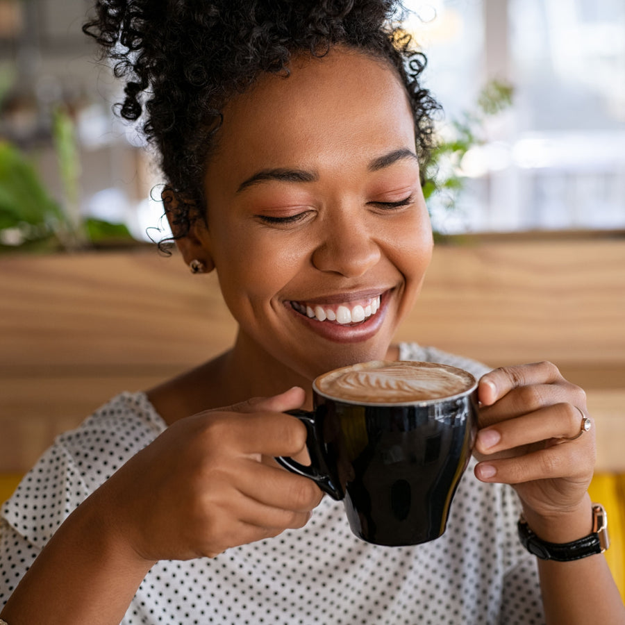A smiling woman with curly hair holds a black coffee cup close to her face, enjoying a moment in a cozy café with plants in the background.