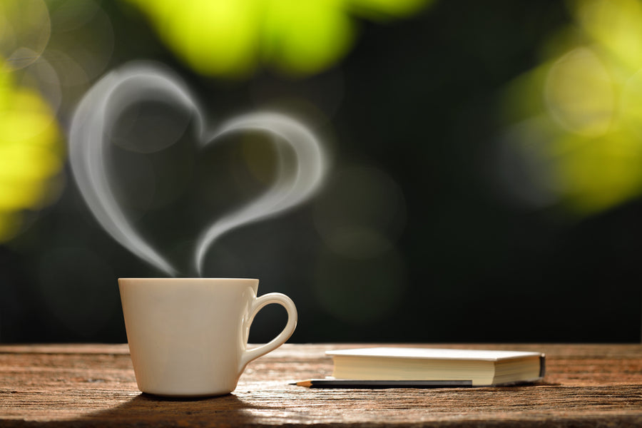 A white coffee cup sits on a wooden table, with steam rising in the shape of a heart, beside an open notebook and pencil, against a softly blurred green background.