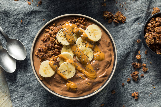 A bowl of chocolate smoothie topped with banana slices, peanut butter drizzle, chia seeds, and granola sits on a textured gray cloth, with two silver spoons nearby and additional granola in a small bowl.
