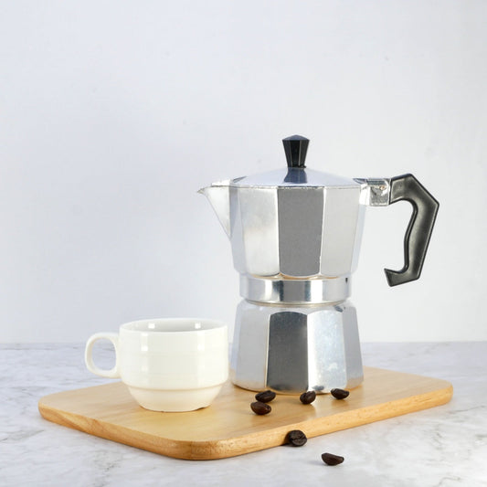 A silver stovetop espresso maker sits on a wooden cutting board next to a white ceramic cup, with a few coffee beans scattered nearby, against a light gray background.