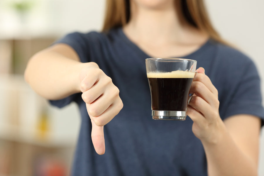 A person in a blue shirt holds a glass of coffee in one hand while giving a thumbs down with the other, expressing disapproval in a bright indoor setting.