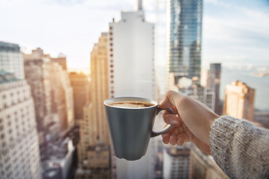 A hand holds a gray coffee mug against a backdrop of tall city buildings and a sunset sky, suggesting a moment of relaxation or enjoyment in an urban setting.