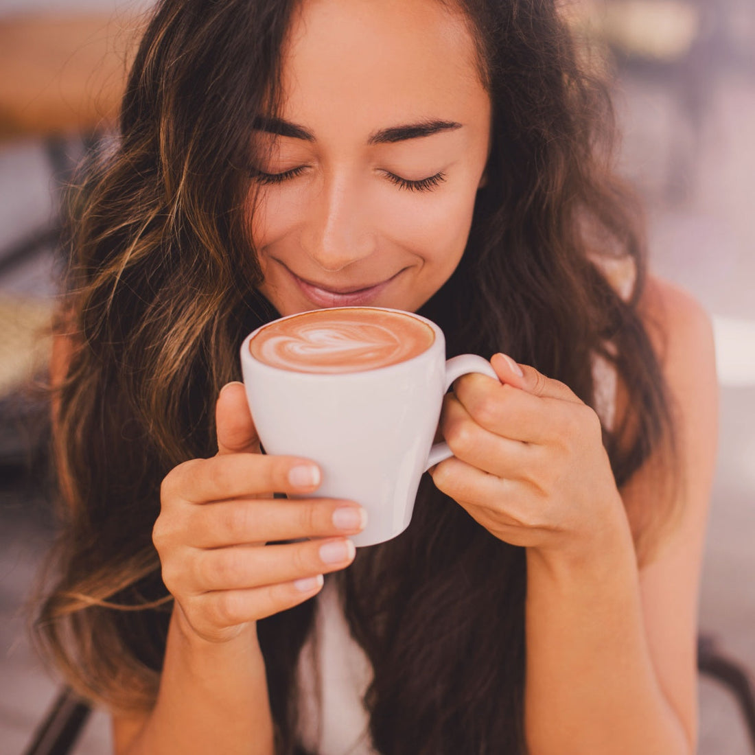 A woman with long, wavy hair smiles gently while holding a white cup of coffee close to her face, enjoying the aroma in a cozy café setting.