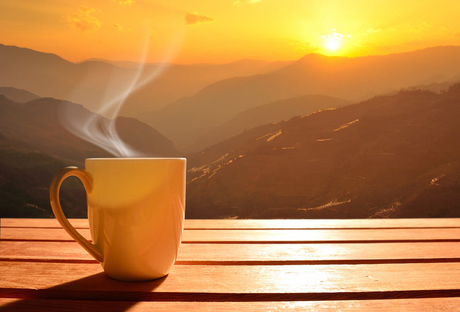 A steaming cup of coffee sits on a wooden table, with a breathtaking view of mountains and a vibrant orange sunset in the background.