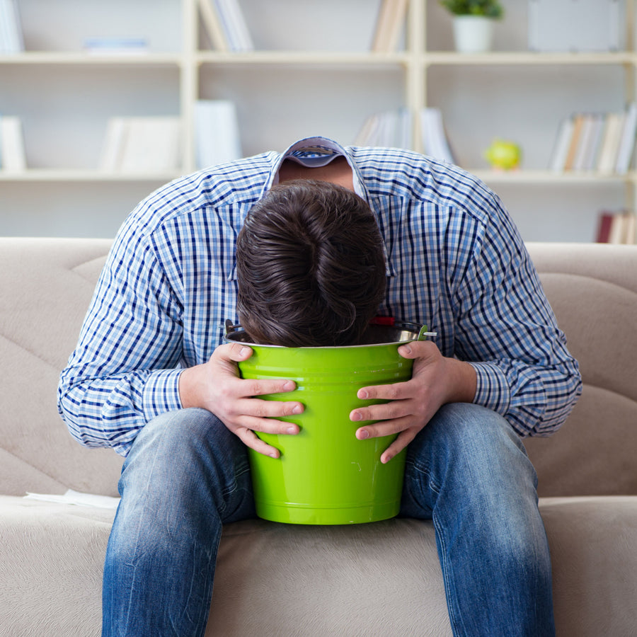 A man in a plaid shirt sits on a couch with his head resting in a green bucket, holding it with both hands, appearing distressed in a cozy indoor setting with bookshelves in the background.