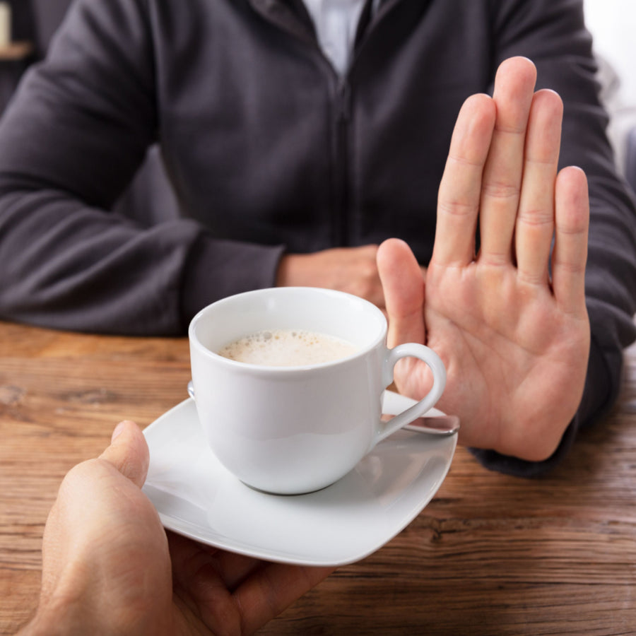 A person is offering a white coffee cup on a saucer to another person, who is raising their hand in a gesture of refusal, set against a wooden table background.