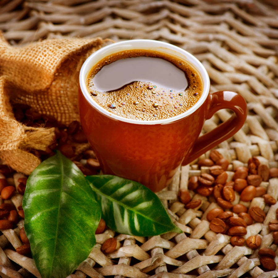 A brown cup filled with dark coffee sits on a woven surface, surrounded by coffee beans and green leaves, with a burlap sack partially visible in the background.