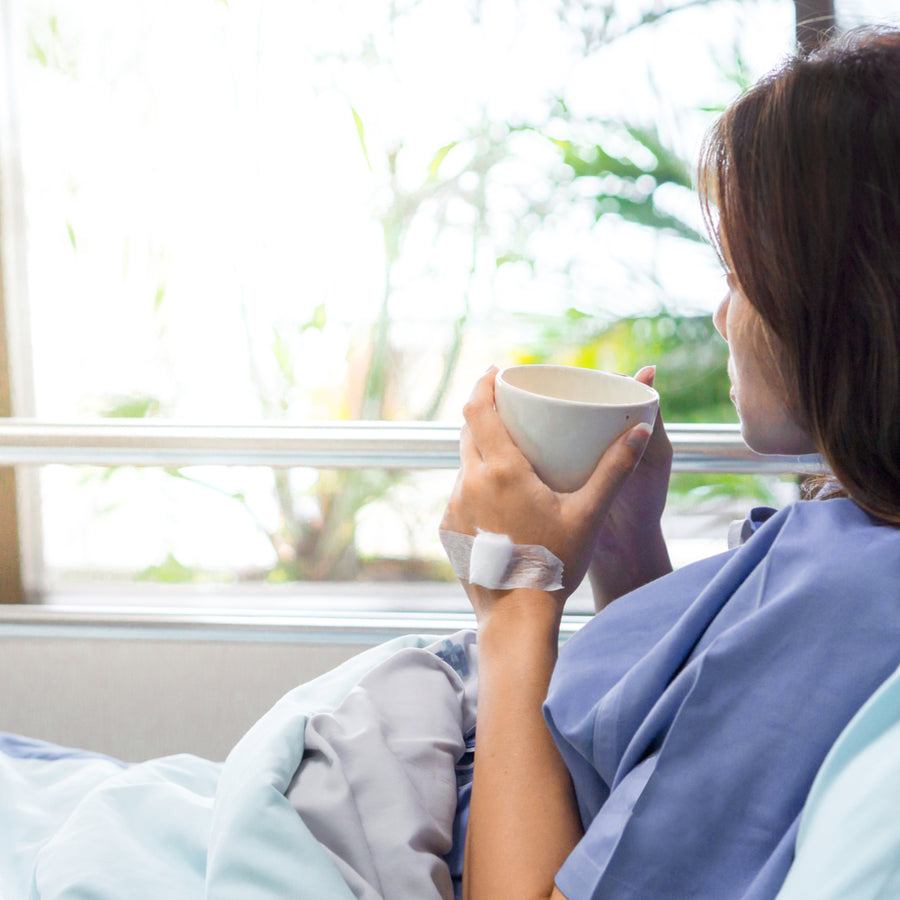 A woman in a hospital bed, wearing a blue gown, holds a white cup while looking out a window with greenery, suggesting a moment of reflection or comfort during recovery.