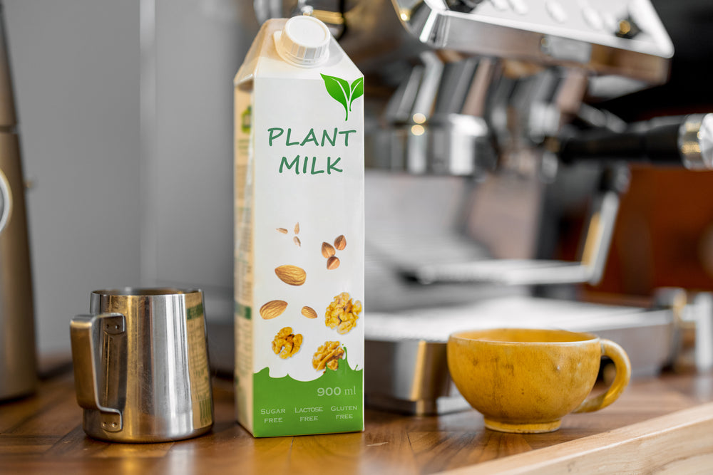 A carton of plant milk labeled "900 ml" stands next to a stainless steel milk frother and a small yellow cup on a wooden counter, with a coffee machine in the background.