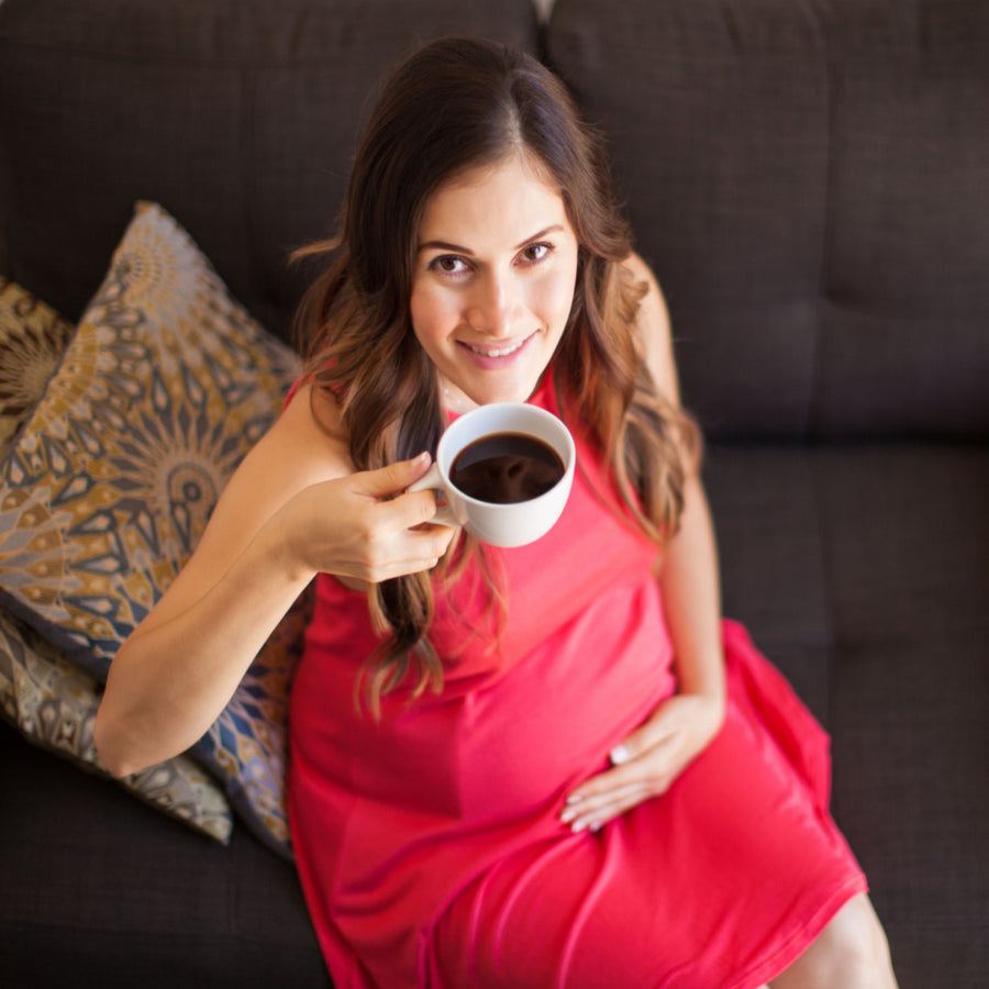 A smiling pregnant woman in a red dress sits on a couch, holding a white cup of coffee while resting her hand on her belly, surrounded by decorative pillows.