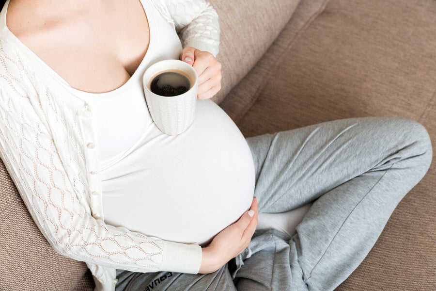A pregnant person sits comfortably on a couch, holding a coffee cup in one hand while gently cradling their belly with the other, wearing a cozy outfit.