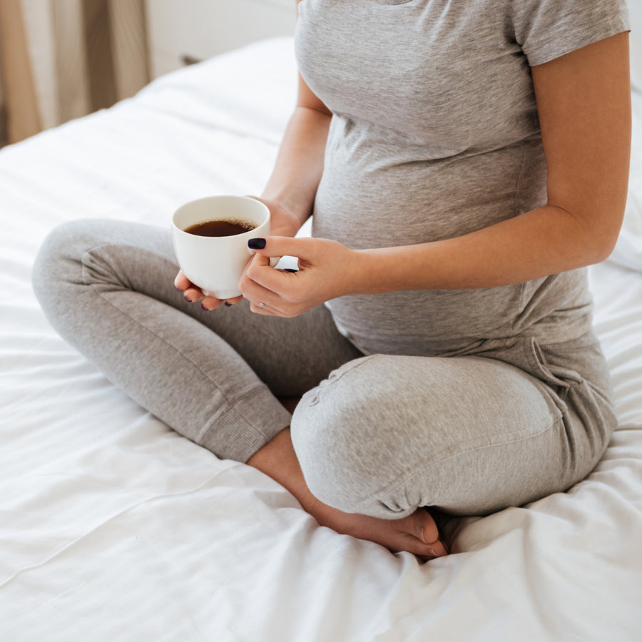 A pregnant person sits cross-legged on a bed, holding a white cup of tea or coffee, wearing a gray outfit, with a calm expression in a cozy, softly lit room.