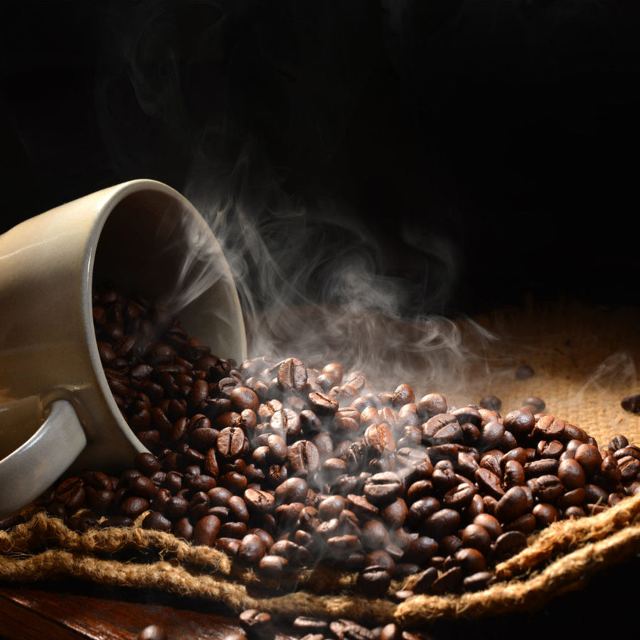A tipped-over beige coffee cup spills aromatic coffee beans onto a textured burlap surface, with wisps of steam rising in a dark setting.