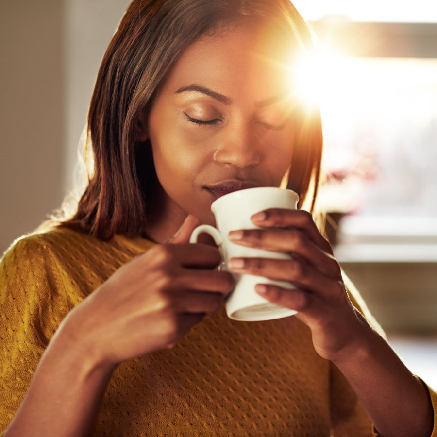 A woman with long hair in a yellow sweater sips from a white mug, eyes closed and a serene expression, as sunlight streams in from a nearby window, creating a warm atmosphere.