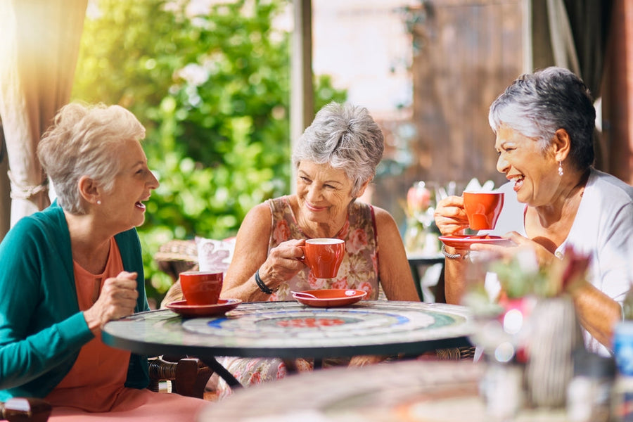 Three older women are sitting at a table in a bright café, laughing and enjoying cups of tea, surrounded by greenery and a warm, inviting atmosphere.