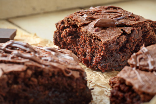 Three rich, chocolate brownies are displayed on crinkled parchment paper, with one brownie partially cut to reveal a moist interior, and drizzled with melted chocolate on top.