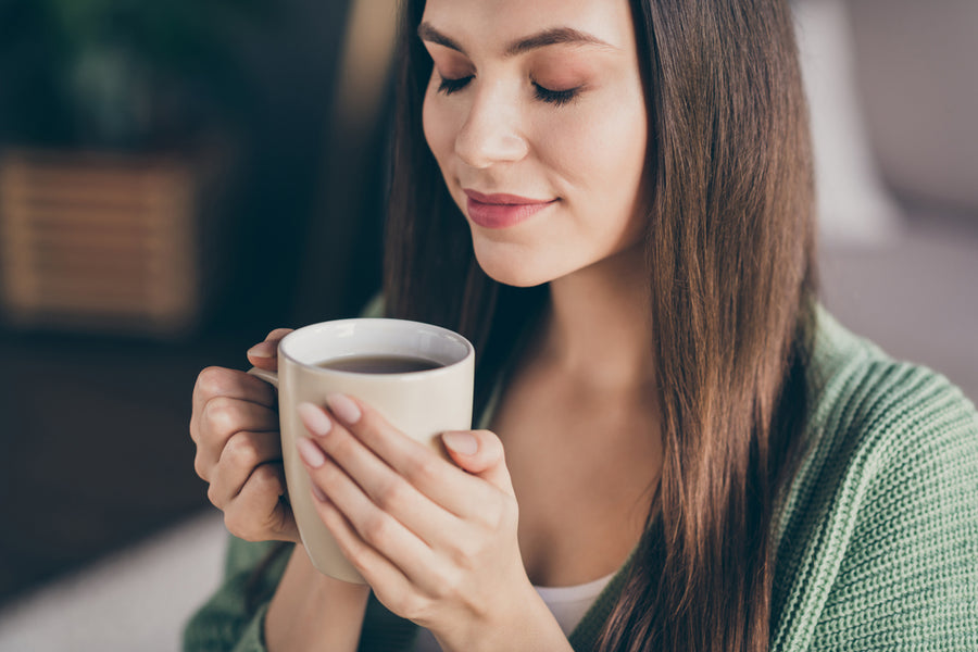 A woman with long hair gently holds a mug of coffee, eyes closed and smiling softly, sitting in a cozy, softly lit room with a green sweater draped over her shoulders.