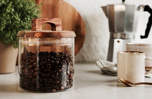 A glass jar filled with dark coffee beans sits on a kitchen counter beside a moka pot, a beige mug, and a stack of napkins, with a green plant in the background.