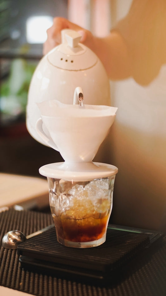 A person pours hot water from a white kettle into a coffee filter resting on a glass filled with ice and dark liquid, set on a black mat in a cozy café environment.