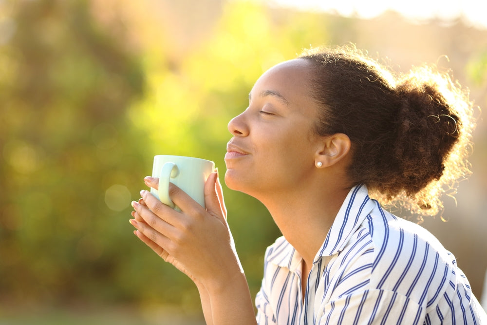 A woman with curly hair enjoys a warm drink, closing her eyes and smiling softly, while sitting outdoors surrounded by a lush, blurred green background.