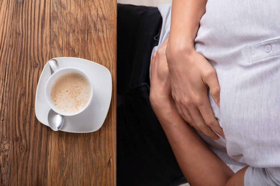 A person sitting at a wooden table holds their stomach with one hand while a cup of frothy coffee rests beside them, suggesting discomfort or contemplation.