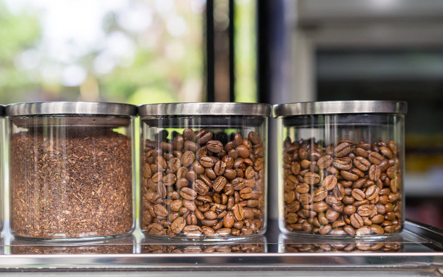 Three glass jars with metal lids are filled with different coffee products: ground coffee on the left, roasted coffee beans in the middle, and another type of roasted beans on the right, set against a blurred outdoor background.