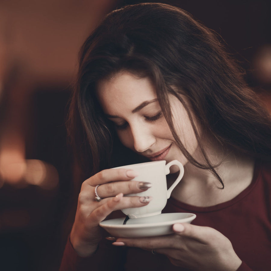 A woman with long hair gently holds a white teacup close to her face, smiling softly as she enjoys the aroma, with a warm, cozy background.