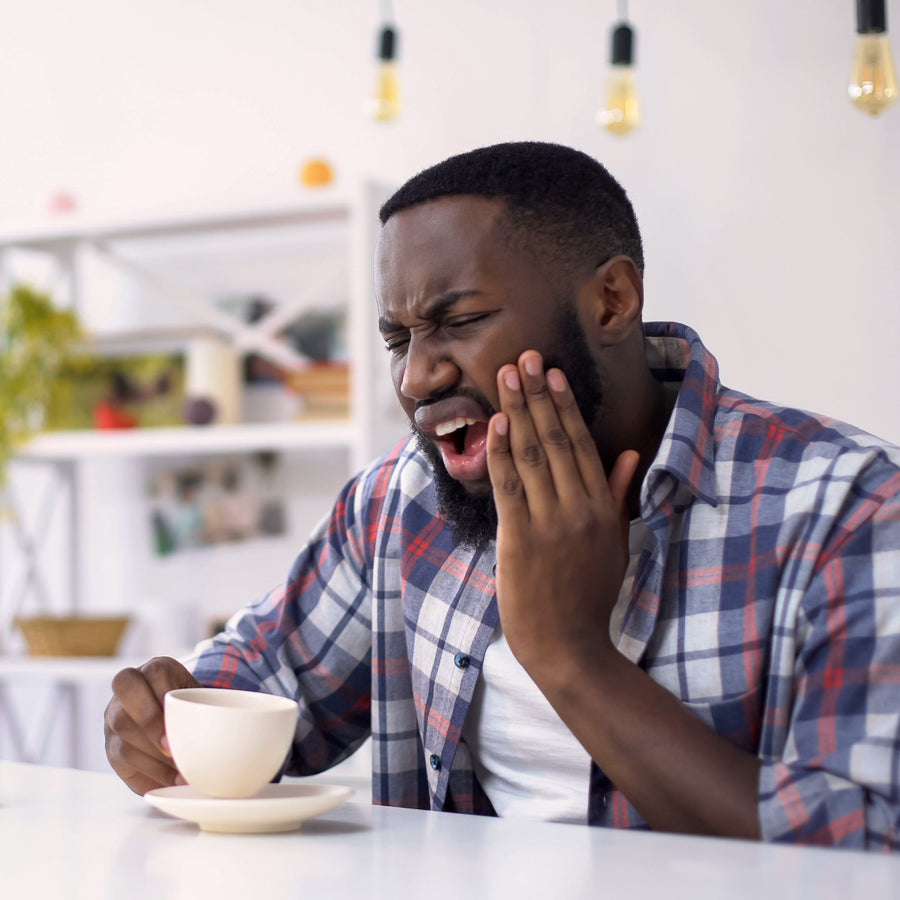 A man in a plaid shirt grimaces in pain while holding a cup, sitting at a table in a bright, modern café, suggesting he is experiencing a toothache.